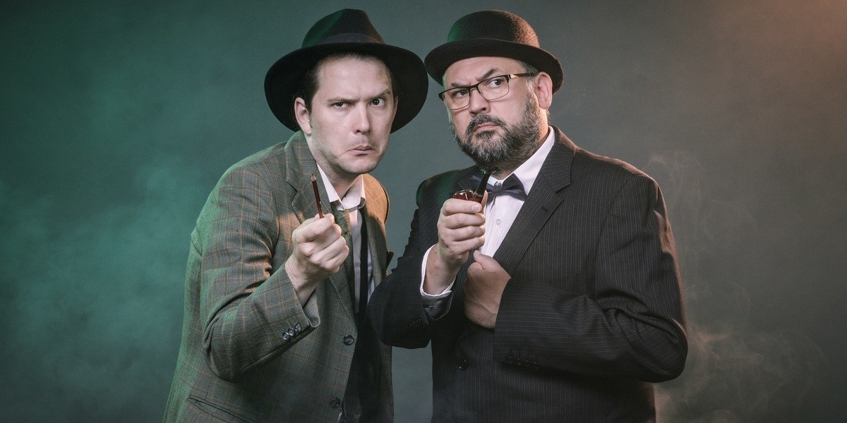 Murder Village: An Improvised Whodunnit - Two men dressed in mid-20th century suits stand in a foggy environment and look off beyond the camera mysteriously. The man on the left wears a fedora and holds a notepad and pencil, while the one on the right has a bowler hat and a smoking pipe.