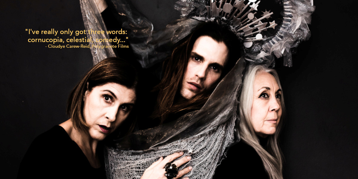 Two women stand cradling a man either side of him, all wearing blacks. The woman on the left has dark honey hair to her chin. The woman on the right has long, grey hair. The man in the middle has shoulder length brown hair and is wearing a grey bejewelled Crown with netting on it with a circular piece on the top. There is grey netting draped over the front of them.