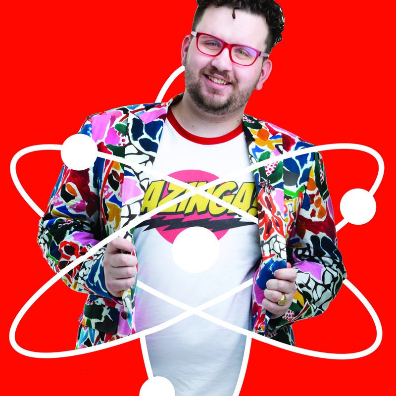 A white man with curly dark hair and facial stubble is smiling. He is wearing a white shirt with Bazinga on the front and a brightly coloured patterned jacket. There is a red background with white lines representing atoms moving around him.