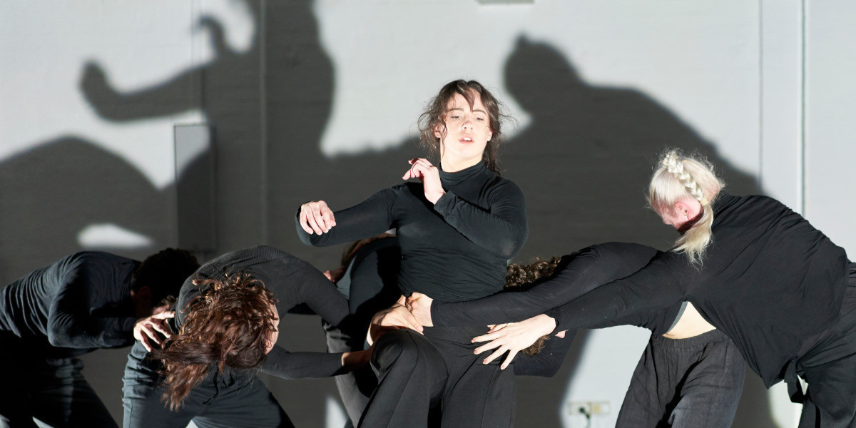A group of dancers are in a mass with a central dancer upright and arms outstretched. The image has a tension as the others are bent over and holding onto the waist of the central dancer. On the back wall the shadows are enlarged and create atmosphere. All the dancers wear black fitted long sleeve tops and bottoms. This is a section created by Lina Limosani from the October 2021 X - available to watch/listen to on YouTube. Photo credit Sam Roberts.