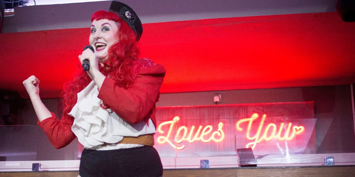 Viva GlasVegas: A Burlesque Showcase from Scotland - Roxy Stardust stands on stage talking into a mic. She has red hair, a red jacket with fringe, shirts and a shirt with frills. She wears a black hat.