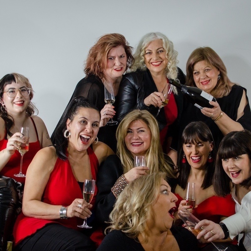 Nine women dressed in red and black clothing holding champagne flutes and smiling and laughing.