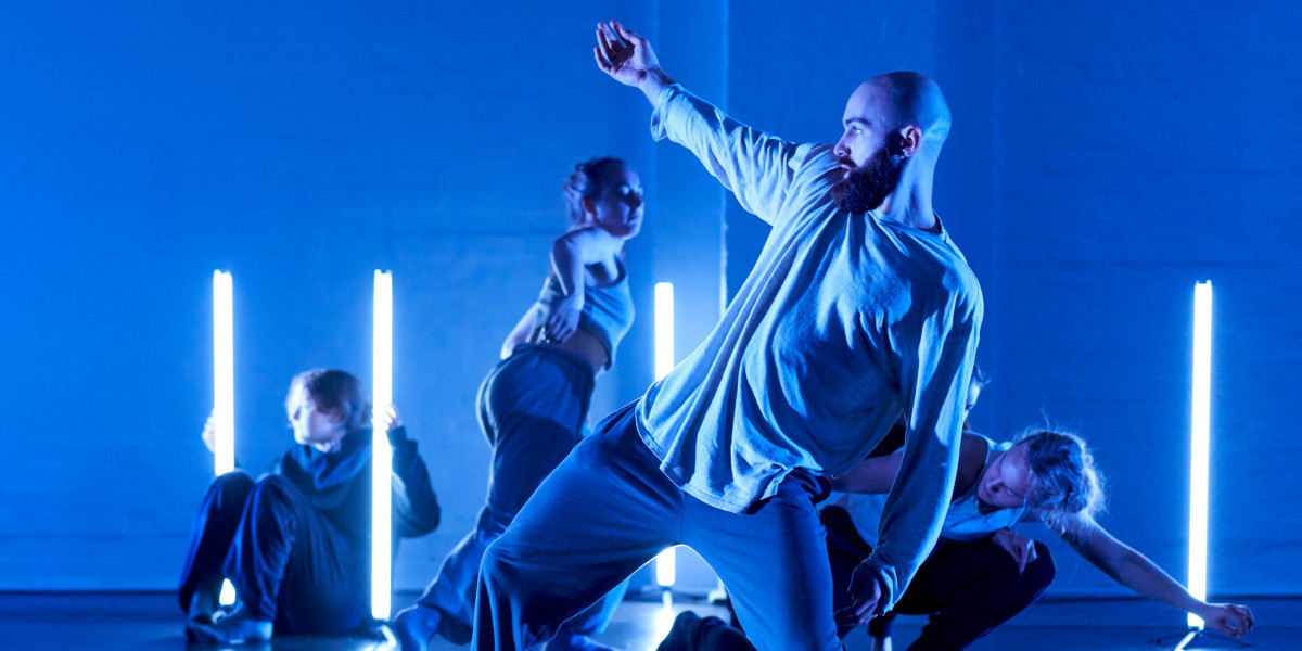 In the foreground is a male dancer lunging. They are side on with one arm outstretched. Three other dancers crouch and lean near white tube lighting in the background. They all wear rehearsal attire. The dancers are bathed in blue light reflected on their skin and the wall that is a backdrop.  This is a section by Lewis Major from August's X available to watch/listen to on YouTube. Photo Credit: Sam Roberts