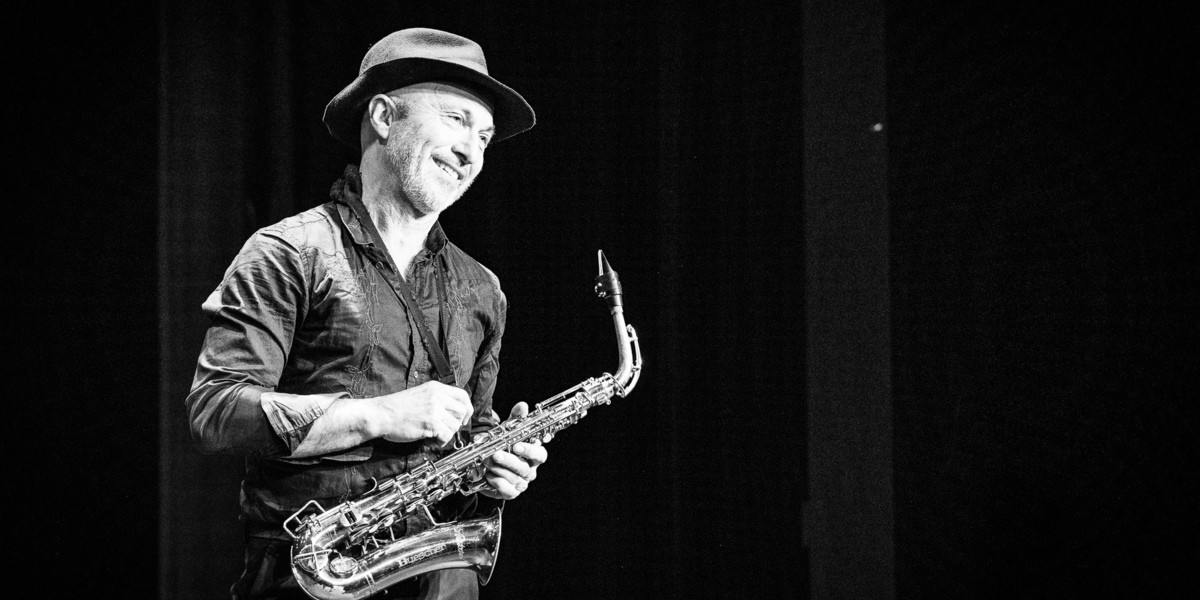 An older man wearing a vintage hat and holding a saxophone smiles.