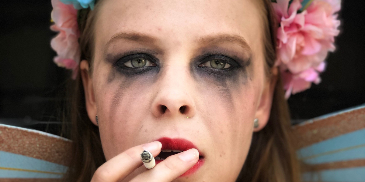 A close up of a womans face, with smeared dark eye make up. She wears a flower crown and smokes a cigarette