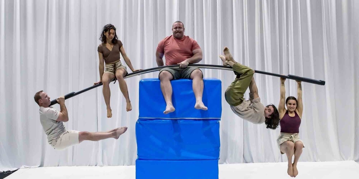 A stack of three large blue blocks, sitting on top is a large man with a long bar across his legs. Hanging from each side of the bar are two acrobats.