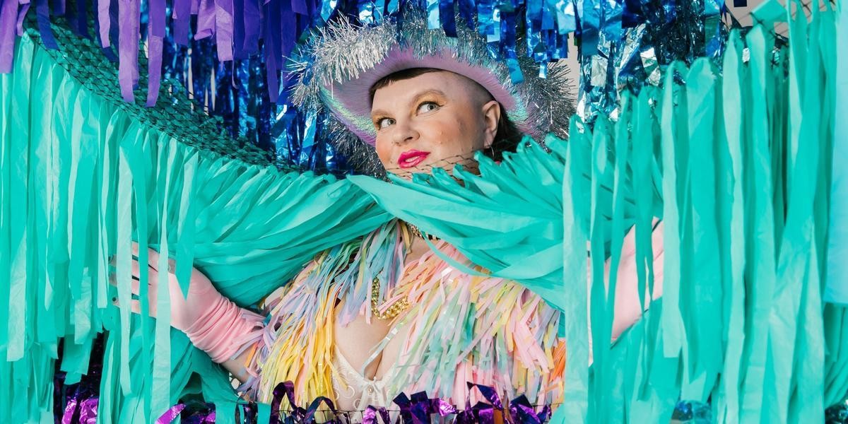 Annie Schofield is posing in a backdrop of blue and green shimmery streamers. They are parting some of the streams so their head sticks out the top. They are wearing a silver Cowgirl hat and a pastel pink tinsel jacket and pink gloves. They are smiling mischievously