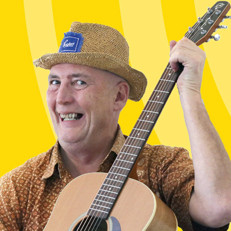 An older man with a large smile wears a straw hat with a blue Coopers Beer logo and a brown patterned shirt. He is holding an acoustic guitar in front of a two toned yellow background.