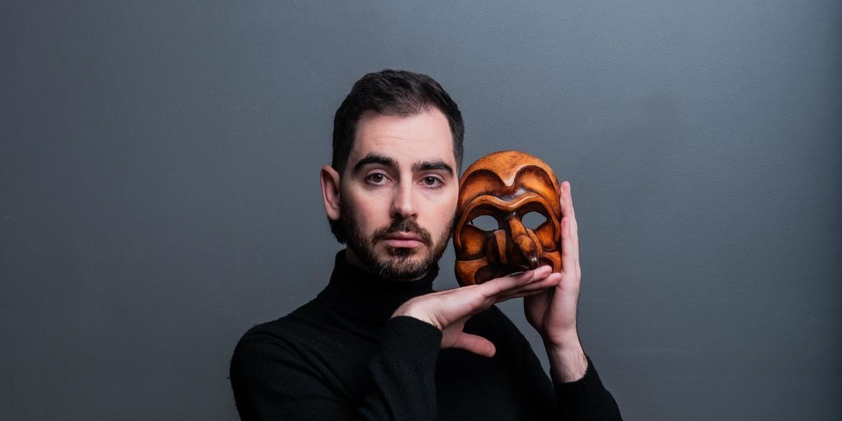 A man in a black turtleneck sweater holds a comedia del'arte mask close to his face with a pretentious and arrogant expression.