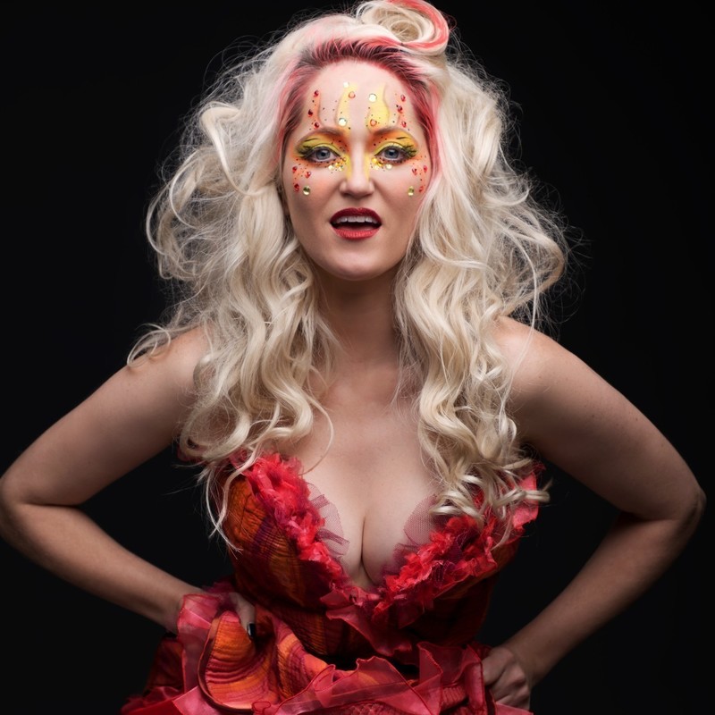 Hotties - A woman with wild long wavy blonde hair, leaning slightly forward with her hands on her hips. She has extravagant yellow, orange and crystal make-up and a low cut ruffled red and orange corset.