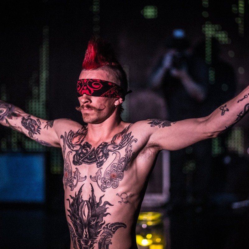 A tattooed man, with no shirt on and a blindfold, holds his arms out. The man has a long, curled moustache and a red mohawk.