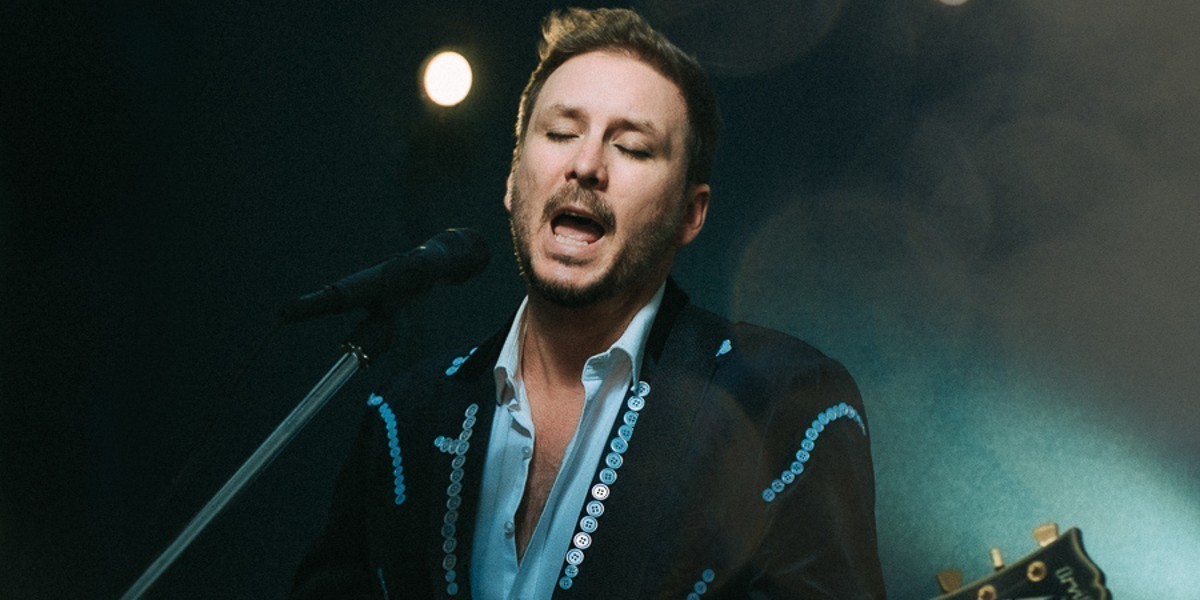 A man playing guitar, singing into a microphone, he is wearing a black jacket