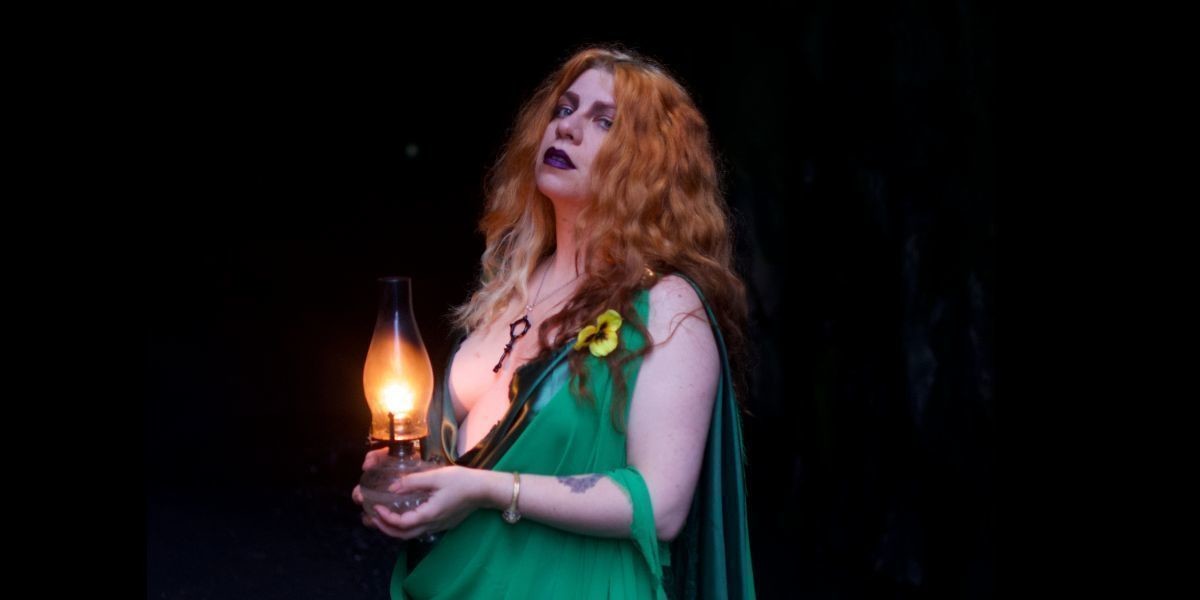 A white women with red curly hair wears a draped green gown stand with her body turned to the right while holding and antique oil lamp in both hands t her stomach while making eye contact with the viewer.