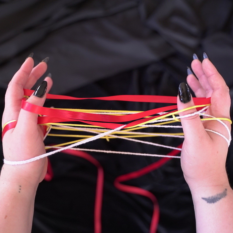 Stuck - The image is of two hands on a black background. The hands are facing each other palm to palm and are stuck in a Cat's Cradle position with interlacing red ribbon, white twine and yellow string.