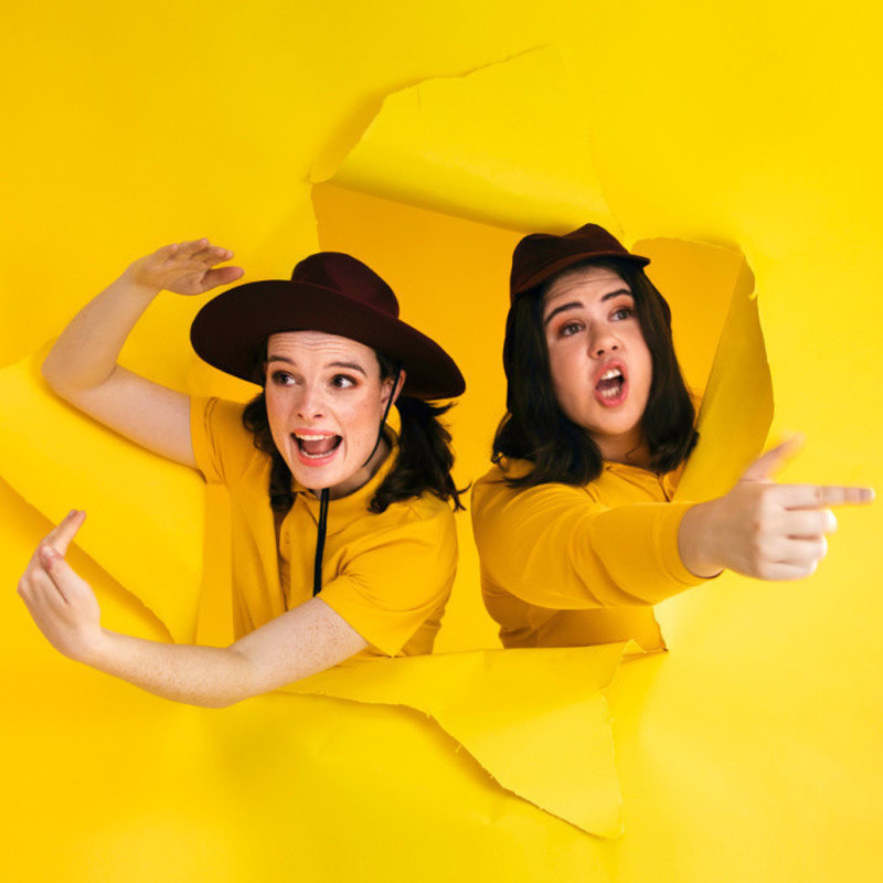 Two white women wearing bright yellow polo shirts and burgundy schools hats burst through a torn piece of yellow cardboard that takes up the whole image. They beckon with enthusiastic facial expressions to the audience to come and join them.