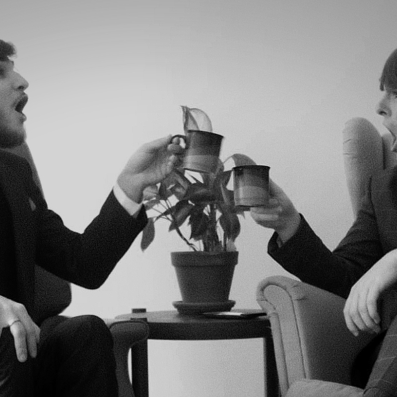 Black and white photograph of Aiden Willcox and Isaac Haigh sitting down in armchairs, pulling silly faces and clinking their mugs together