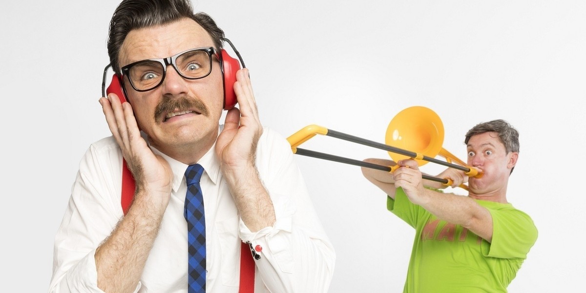 One artists looks down the camera with a pained expression on their face, they are wearing large red headphones, a white shirt and glasses. In the background another artist in a green shirt is blowing a toy trombone in their direction.