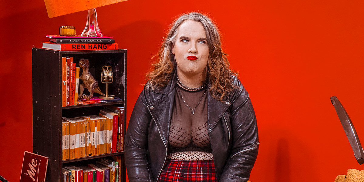 Anna sits down sight a grumpy face and pursed lips, she's wearing fishnet and leather, the background is bright coral red, with a bookshelf and orange books and props