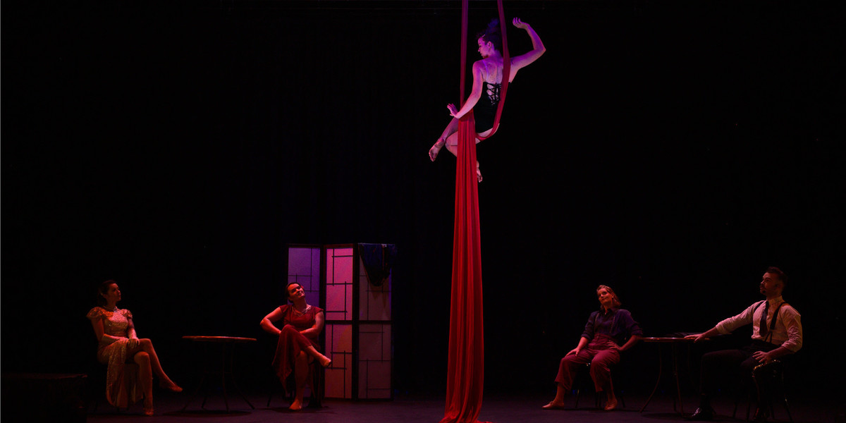 A performer sits high above the stage wrapped in a red silk-like circus apparatus. Below her performers in vintage costumes sit at cabaret tables.