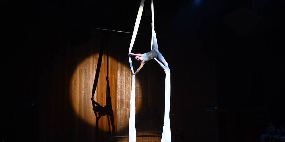 Teenager is doing the splits in the air on a white aerial silks, there is a shadow on the brown wall behind them