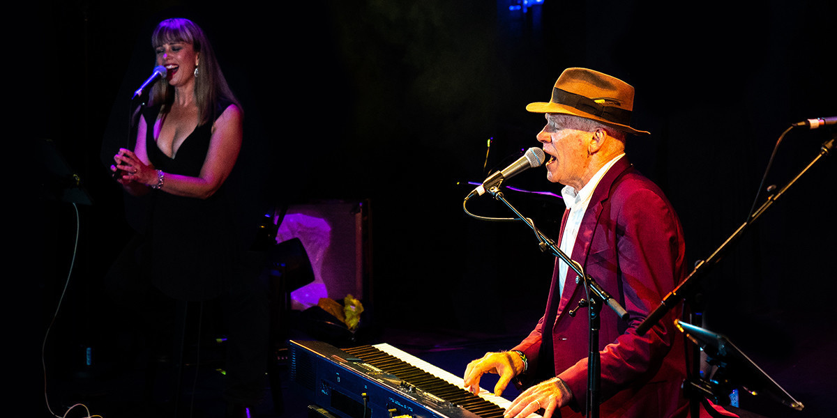 Stewart D'Arrietta on piano roars out a Leonard Cohen song whilst backing singer Emily Kelly accompanies him during a live performance. They are both facing left, and Stewart has both hands on the keyboard, whilst singing into the mic and giving the song his all. He's wearing a burgundy Paul Smith suit, crisp white shirt and his signature hat.
Emily is standing behind him, also singing full pelt into a standing mic, whilst clapping her hands enthusiastically at the same time. She has long blonde hair, sparkly earrings, and is wearing a low cut little black dress.