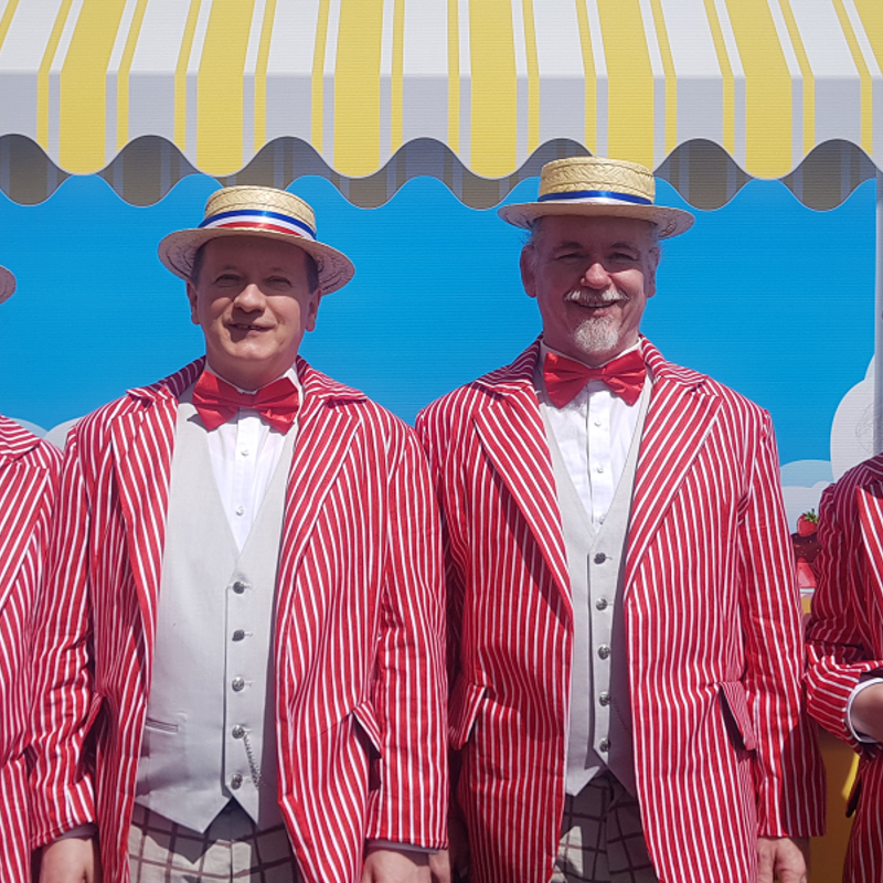 4 People standing in a row all wearing the same costume; a boater hat, a red bow tie, white shirt, silver grey waistcoat and a red stripy jacket standing in front of the picture of an ice-cream cart.