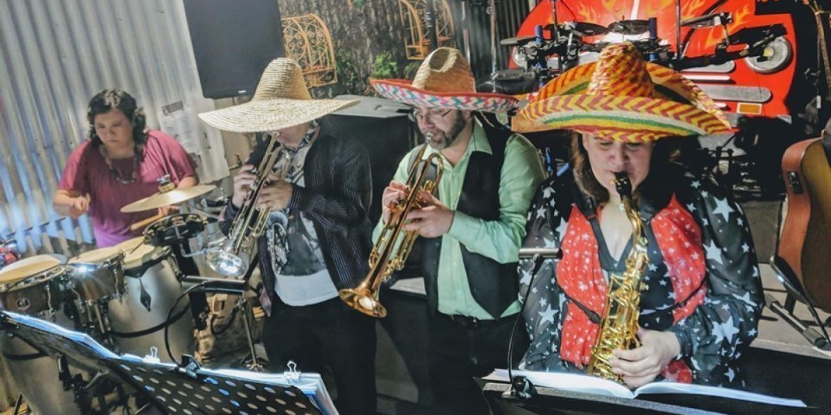 Four musicians, one on percussion, two on trumpets and one on saxophone, with the brass players wearing huge colourful sombreros. All of them are looking at sheet music while the play.