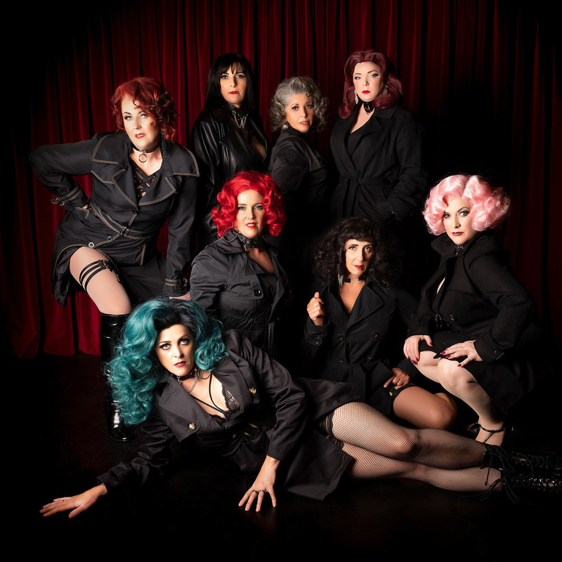 8 women dressed up in evening hair and makeup, wearing black trenchcoats with glimpses of stockings.