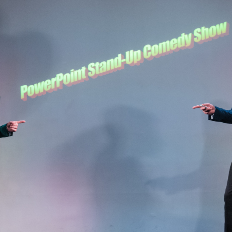 Two men in suits and bowties stand in front of a large screen that reads PowerPoint Stand Up Comedy Show.