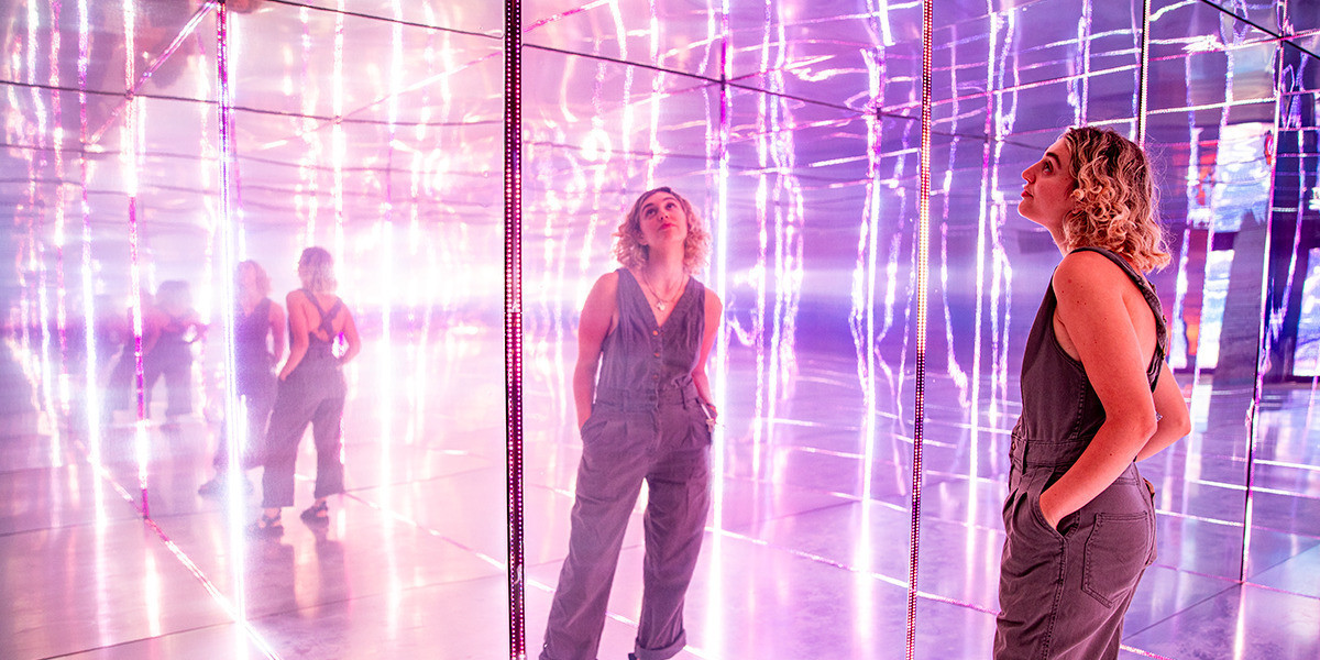 A woman standing with her hands in her pockets looking up at a full wall mirror reflecting a room of mirrors and lights
