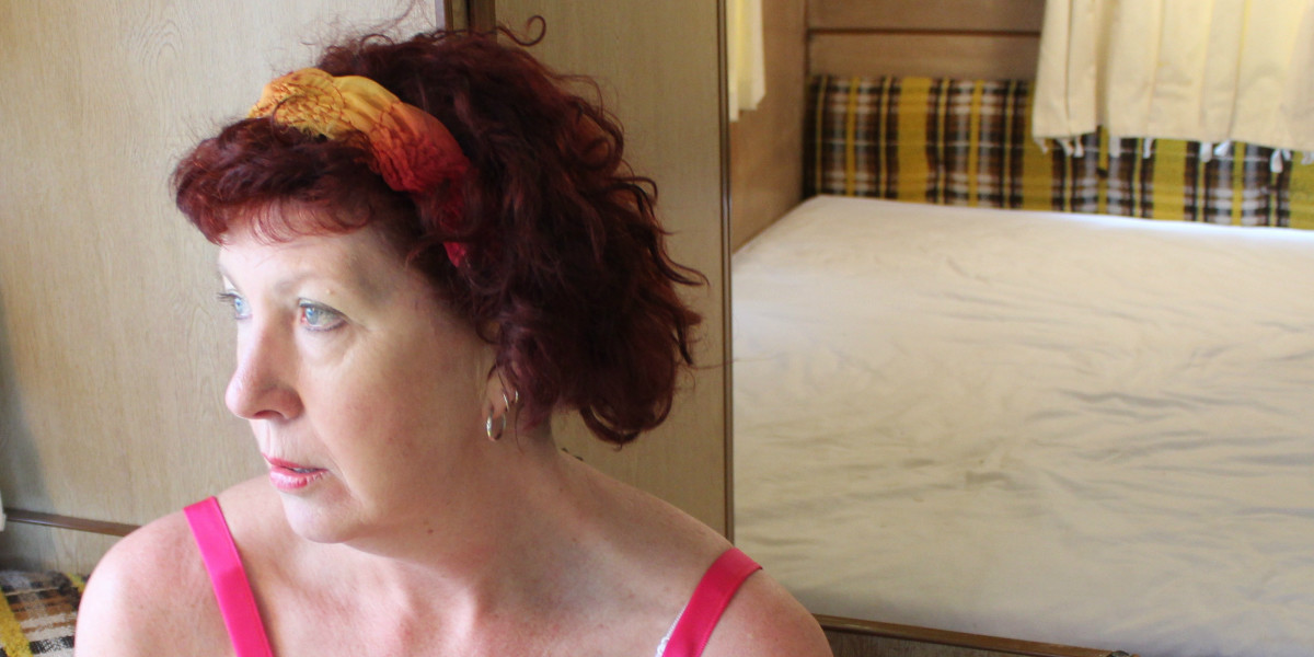 A woman looks pensively to her right with light falling on her face as though she is looking out a window. There are dark pink straps over her shoulders and she wears a pink and yellow headband in her short red curly hair. In another room behind her is a bed with a white sheet on it. The wall behind it is brown and yellow checked  material and a cream curtain covers a window.