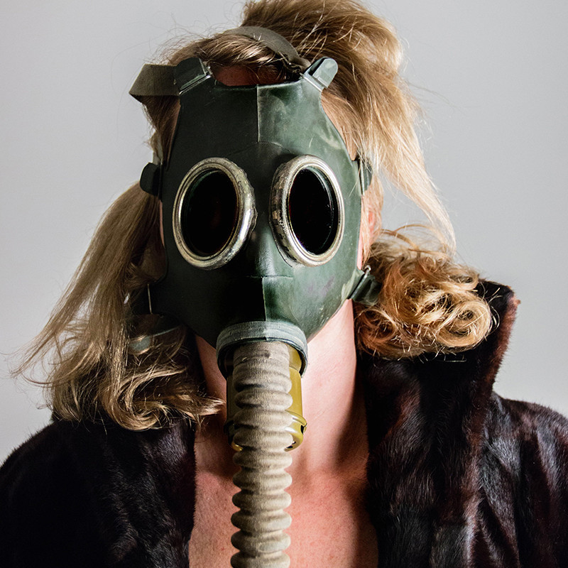 A blond woman's entire face is covered a dark green WWII gas mask. She has on a brown fur coat.