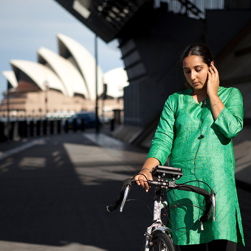 A person standing next to a bike, wearing a longline top, looks down at a smartphone mounted on their bike, with their left hand holding headphones to one ear. The Sydney Opera House appears behind them slightly out of focus.