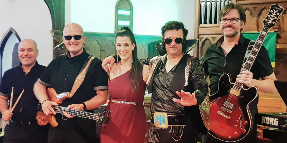 Frank is holding his drum sticks next to Nige who is holding his bass guitar. In the middle Dani has her maroon dress on and Tony is dressed as Elvis Presley. At the end Ken is holding his guitar.