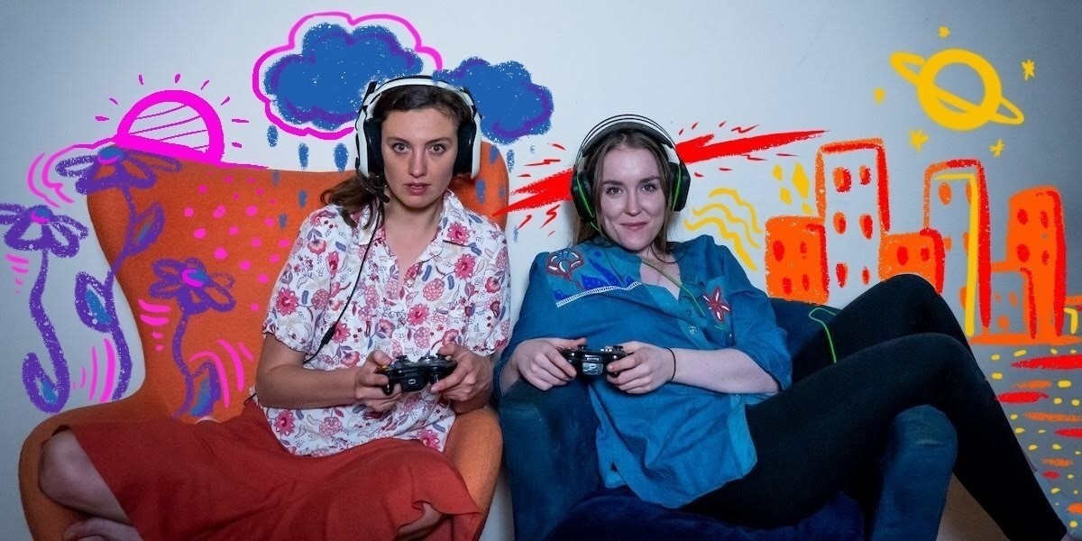 Two young women sit on arm chairs next to each other. They are both holding gaming controllers and look ready for battle. There is colourful graffiti all around them.