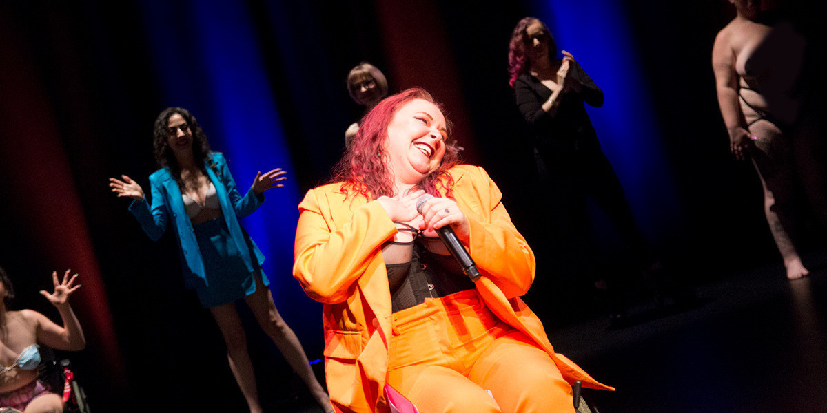 Curtain call at a burlesque show. The host wears an orange suit and sits in their wheelchair laughing in the foreground, with a cast of various performers standing side by side in the background.