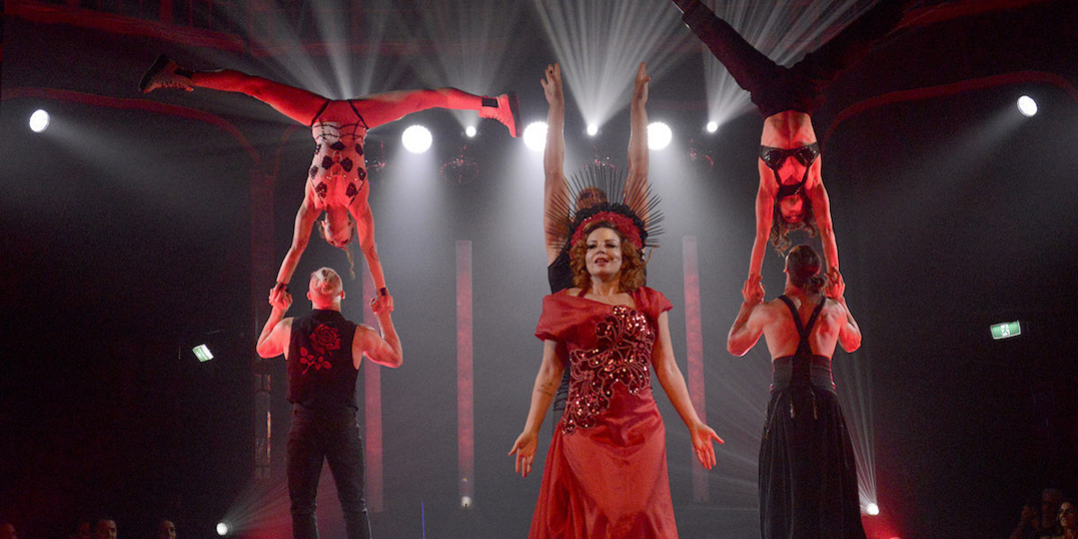 Two pairs either side of the image perform an acrobatic move with an acrobat balancing in a handstand on another person. In the middle a woman with a red gown and elaborate red and black spiky headdress has her arms out