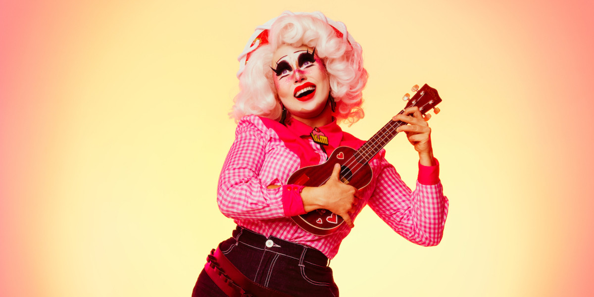 The drag queen Clara Cupcakes is smiling into the distance and holding a ukulele while standing in front of a sunset gradient background. She is dressed as a cowgirl in a red hat with white trim, a pink checked shirt with a pink cupcake bolo tie and denim shorts. Her hair is white and she has a full face of drag make-up.