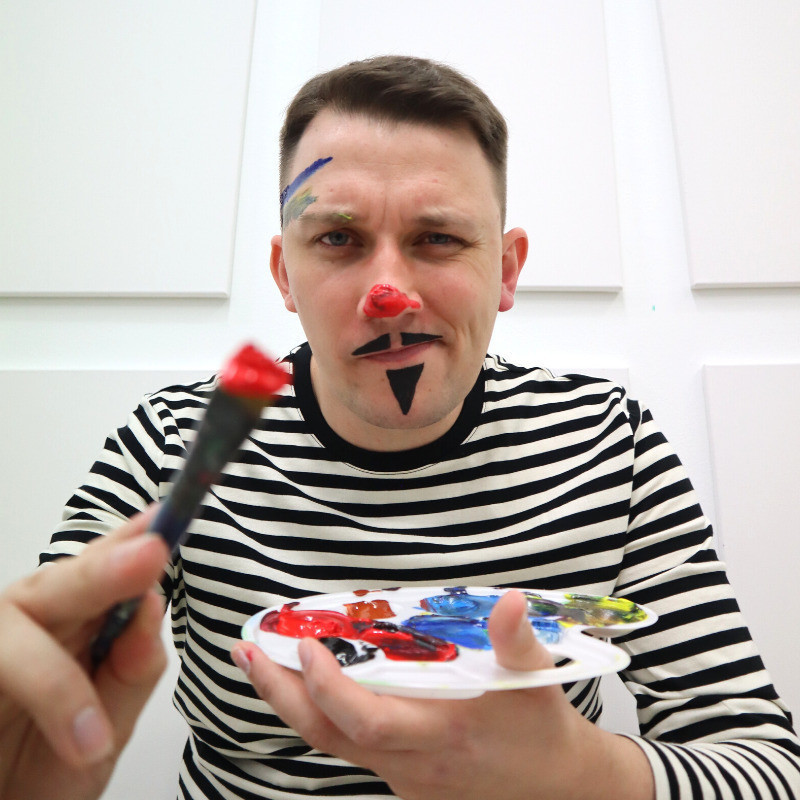 A painter in a black and white striped shirt, with red paint smeared on his nose like a clown, a drawn-on, black triangular moustache and goatee, and blue paint smeared on his forehead, holds an artist's palette and paintbrush loaded with paint as if he is about to paint the camera.