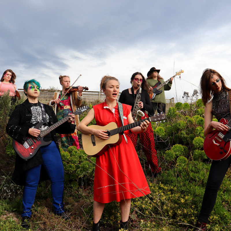 Band playing in a green garden setting, wearing brightly coloured outfits and make up. Each musician has a different instrument: violin, saxophone, 2 guitarist, bass guitar, drums and at the front there's the lead singer in a red dress with an acoustic guitar.