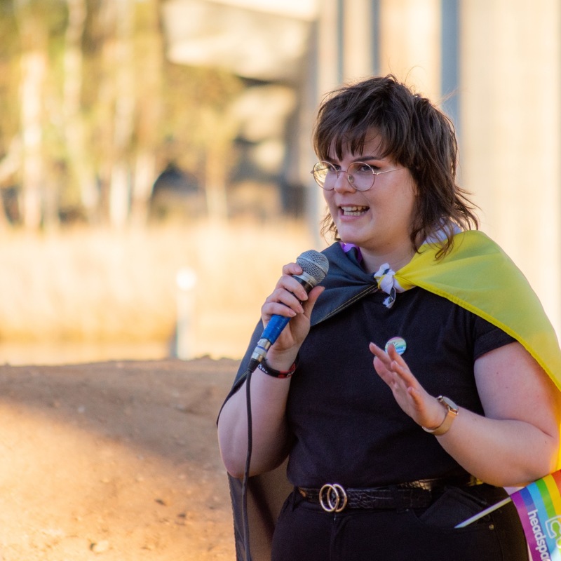 Counter Urban Questions - Non-binary performer Jess speaks into a microphone. They have round glasses and a mullet haircut (with brown hair). They are wearing black clothing, with a non-binary flag as a cape.