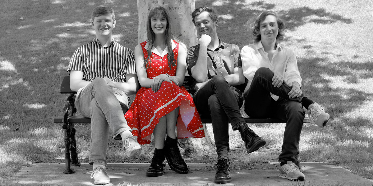 Four performers (Left to Right: Dan Courts, Tiffany Gaze, Jack Barton and Edmund Black) sitting on a garden bench under a tree, all with their legs crossed in a similar fashion. Tiffany wears a red polka-dot dress.