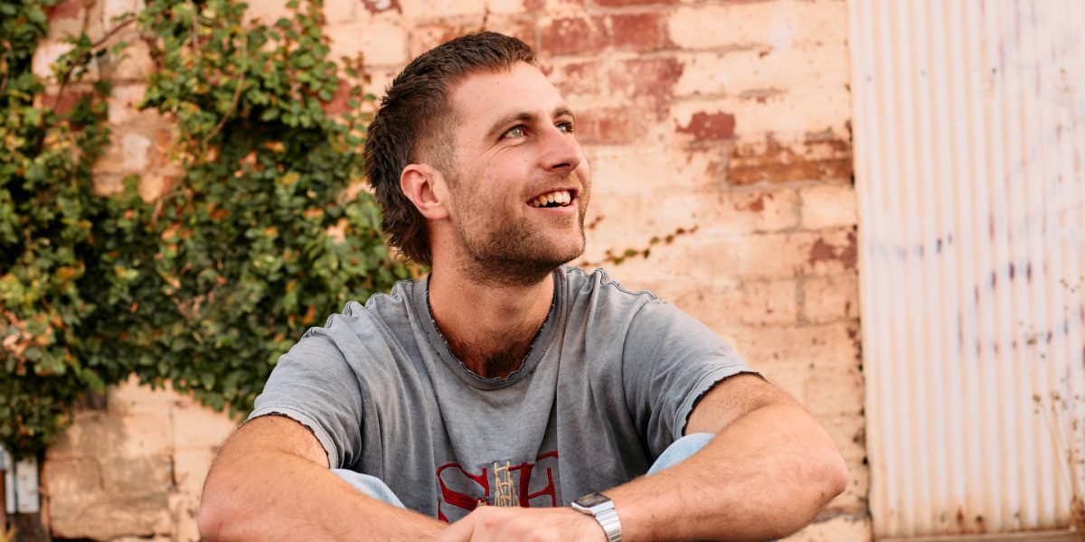 Lewis is sitting outdoors in front of a brick wall. He has short brown hair and is wearing a grey tshirt. He is looking up toward the sky and smiling with his teeth.