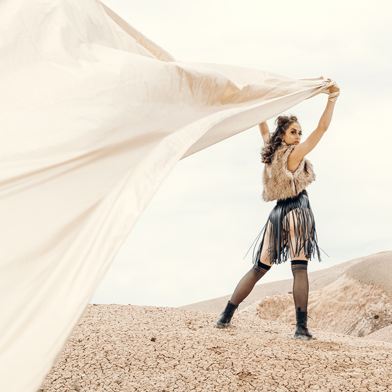 A desert scene with a brown haired woman standing on a hill. She is wearing a fur vest and leather fringed skirt while holding a large gold cape behind her as it blows in the wind.