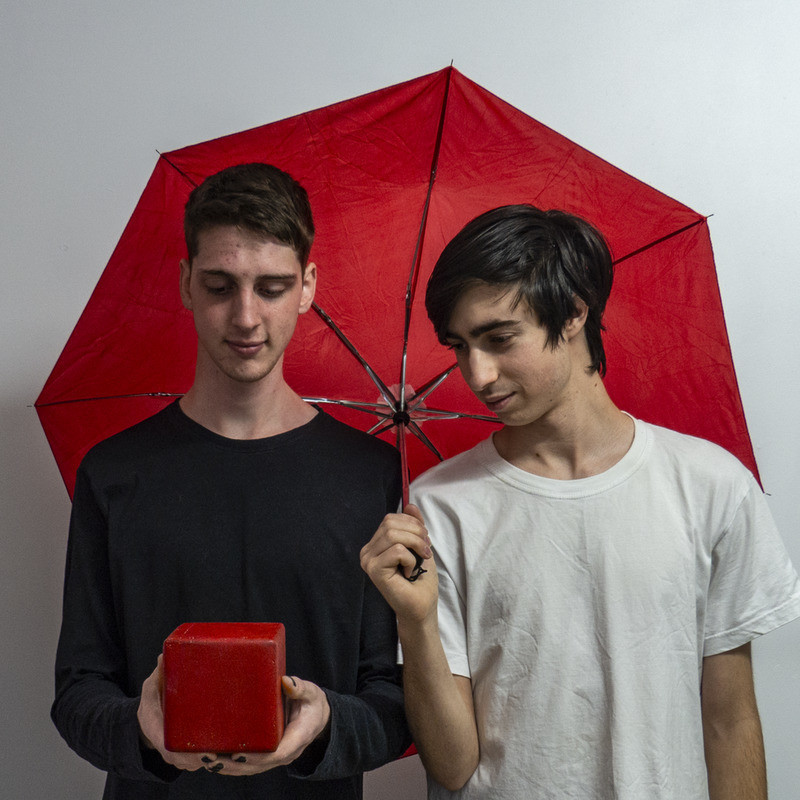 Chroma - A photo of two men looking at a red box. The man on the right is wearing a black long-sleeved shirt and is holding the red box in his hands. The man standing next to him is wearing a white t-shirt and is looking at the box while holding a red umbrella behind his head.