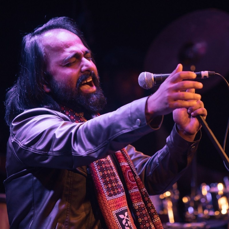 A photo of a man singing passionately into a microphone. He has black hair and a full beard. He is wearing a black leather jacket and an orange, black and white patterned scarf.