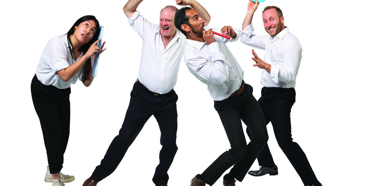 One woman and three men, all in white shirts and black pants stand in a row while dancing each holding a different object