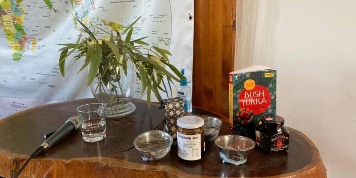 A sprig of eucalyptus leaves stands in a vase on an exposed wood tabletop. Alongside are a range of spices and ingredients, including quandong jam and wild hibiscus. A microphone rests on the table and in the background there is a map of the world.