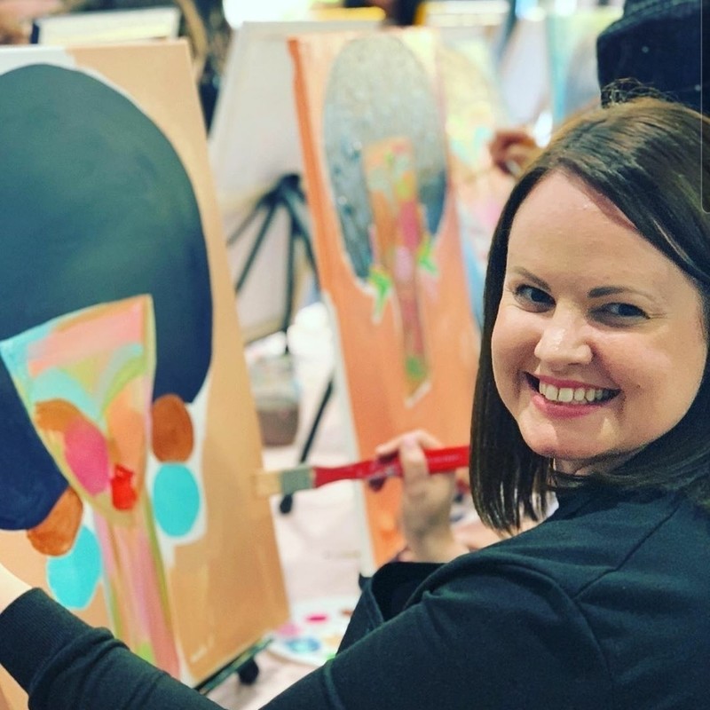 A photo of a woman smiling and holding a paintbrush. The background features the canvas that she is painting on.