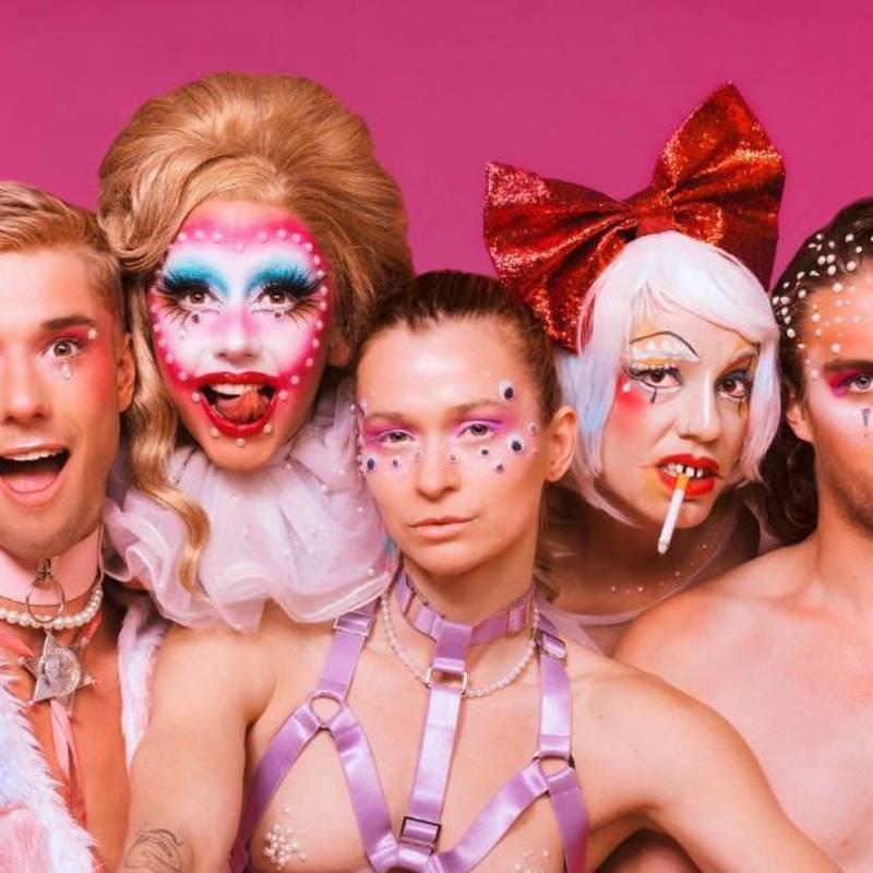 A close up photo of 7 artists from the chest up, all looking into the camera. They are dressed in various costumes in shades of pink and red, most with sparkles on.